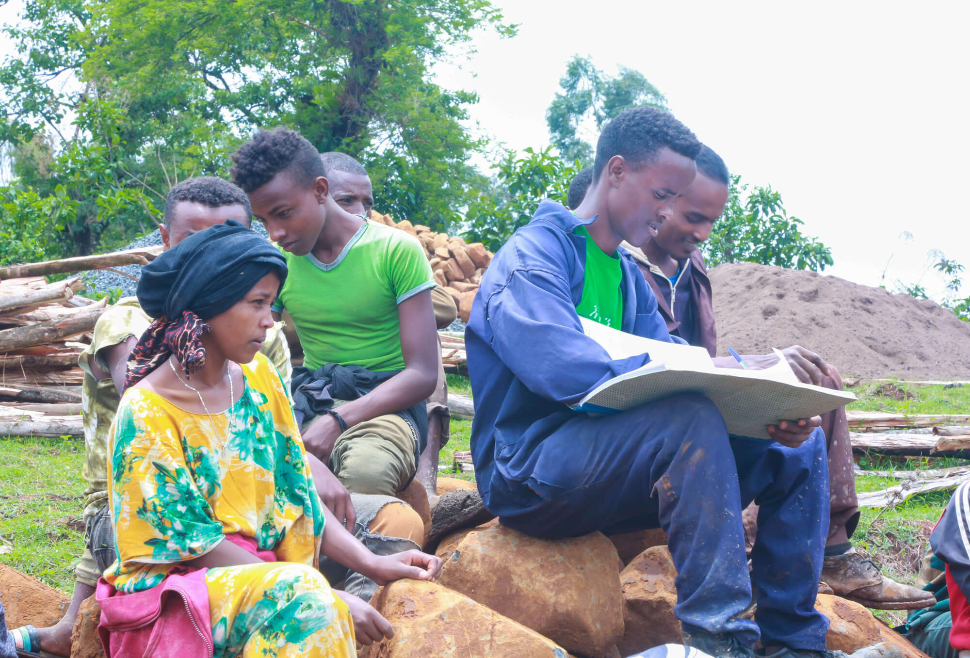 A group of young people sit on rocks, with one person writing in a notebook while others observe intently.