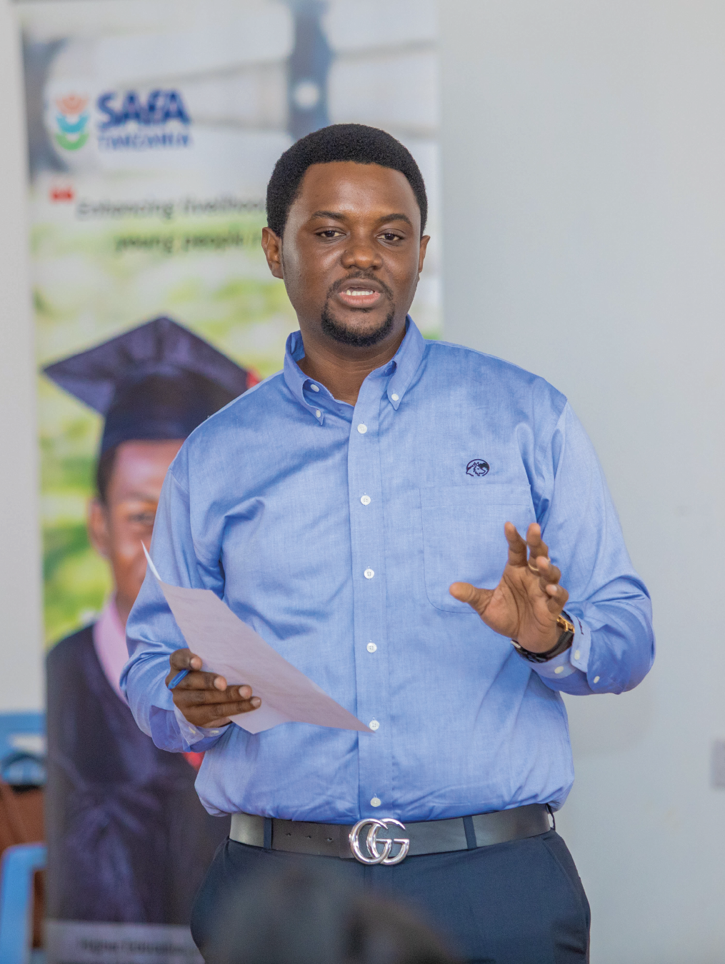 A man in a blue shirt speaks while holding papers, with a promotional banner in the background.