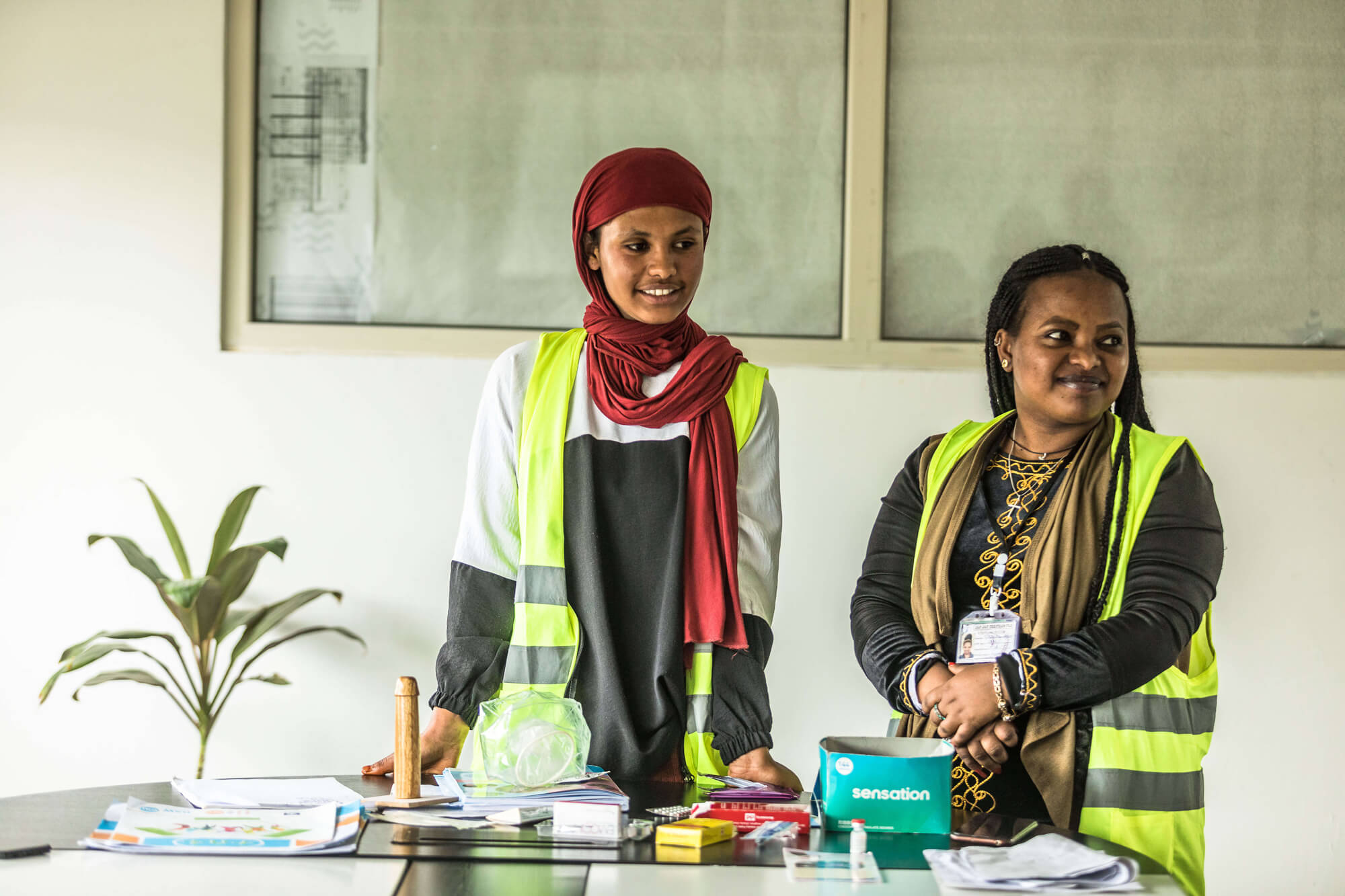 Two women in fluorescent vests stand at a table with documents and supplies, smiling in a bright, modern space.