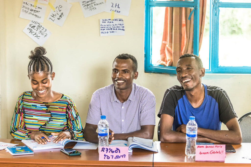 Three individuals sit at a table, smiling and engaged, with notebooks and water bottles in a classroom setting.