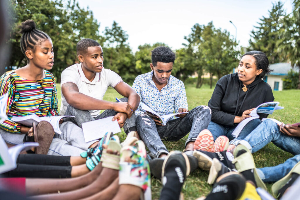 A group of young people sits on grass, engaged in discussion with notebooks and books, focused on their activity.