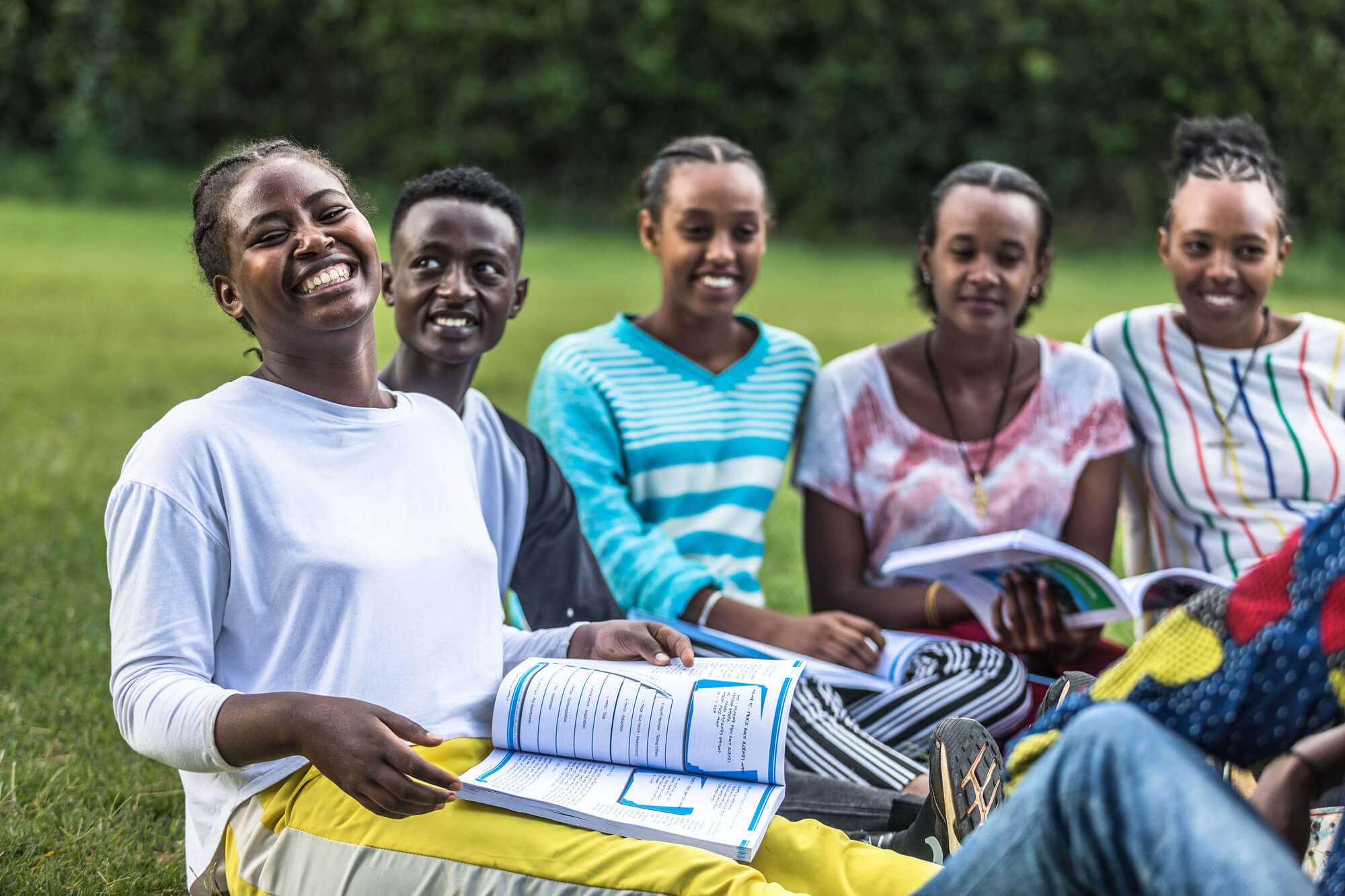 A group of six young people smile while gathered on grass, holding books and enjoying each other's company outdoors.
