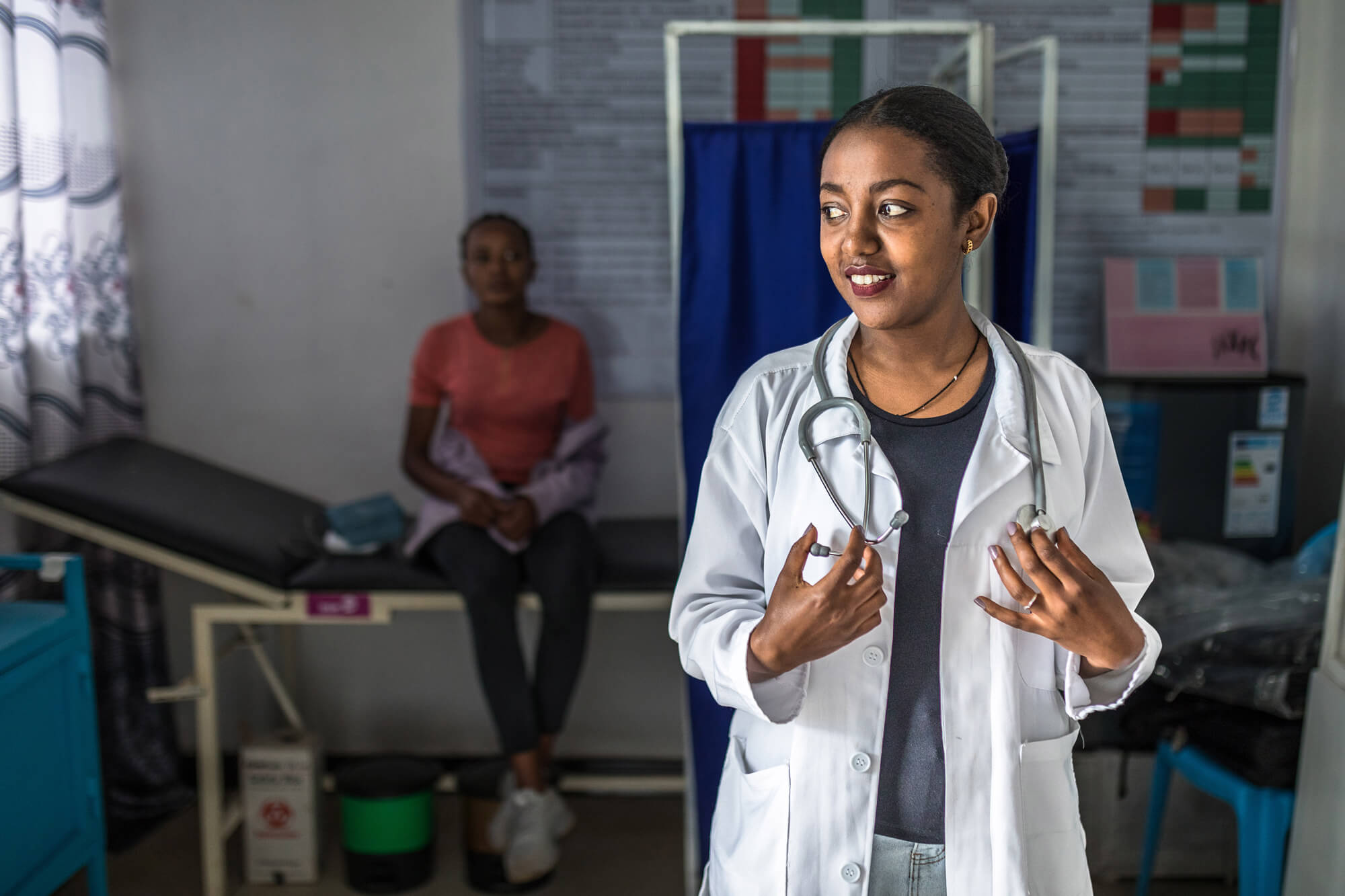 A healthcare professional in a lab coat and stethoscope speaks while a patient sits on an examination table in the backgro...