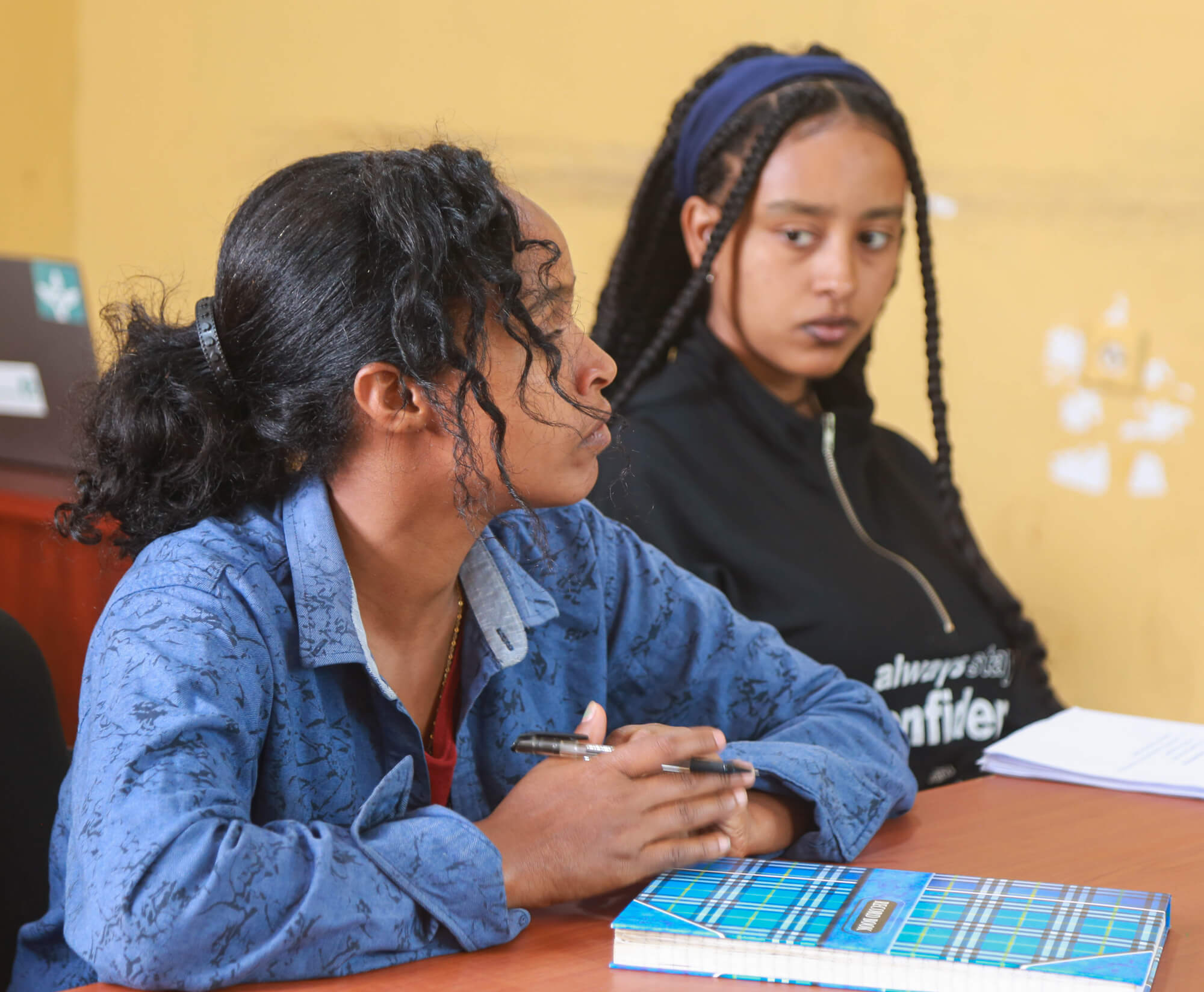 Two young women sit at a table, one with curly hair and the other with long braids, engaged in conversation.