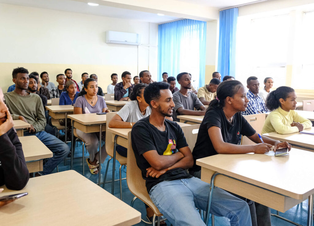 A classroom filled with students attentively listening to a lecture, seated at wooden desks in a well-lit setting.