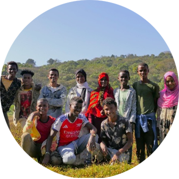 A group of young people stands together outdoors in a grassy field, with hills and clear blue sky in the background.