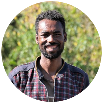A smiling man with a beard, wearing a checkered jacket, stands outdoors with green foliage in the background.