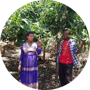 Two individuals stand among lush greenery, one wearing a blue dress and the other in a red shirt, holding fruits.