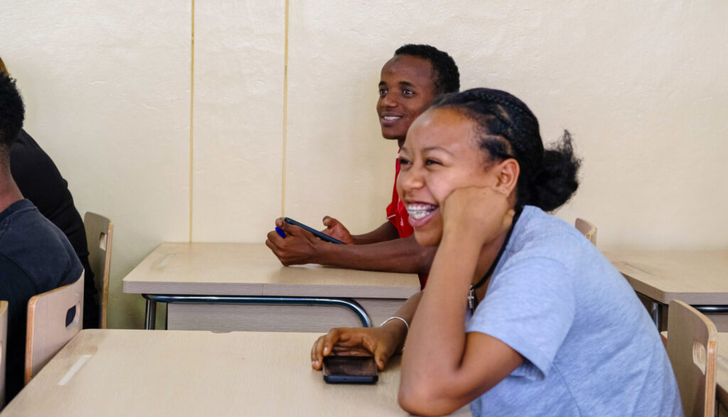 Two young people are sitting at desks in a classroom, smiling and engaged in conversation. One holds a phone.