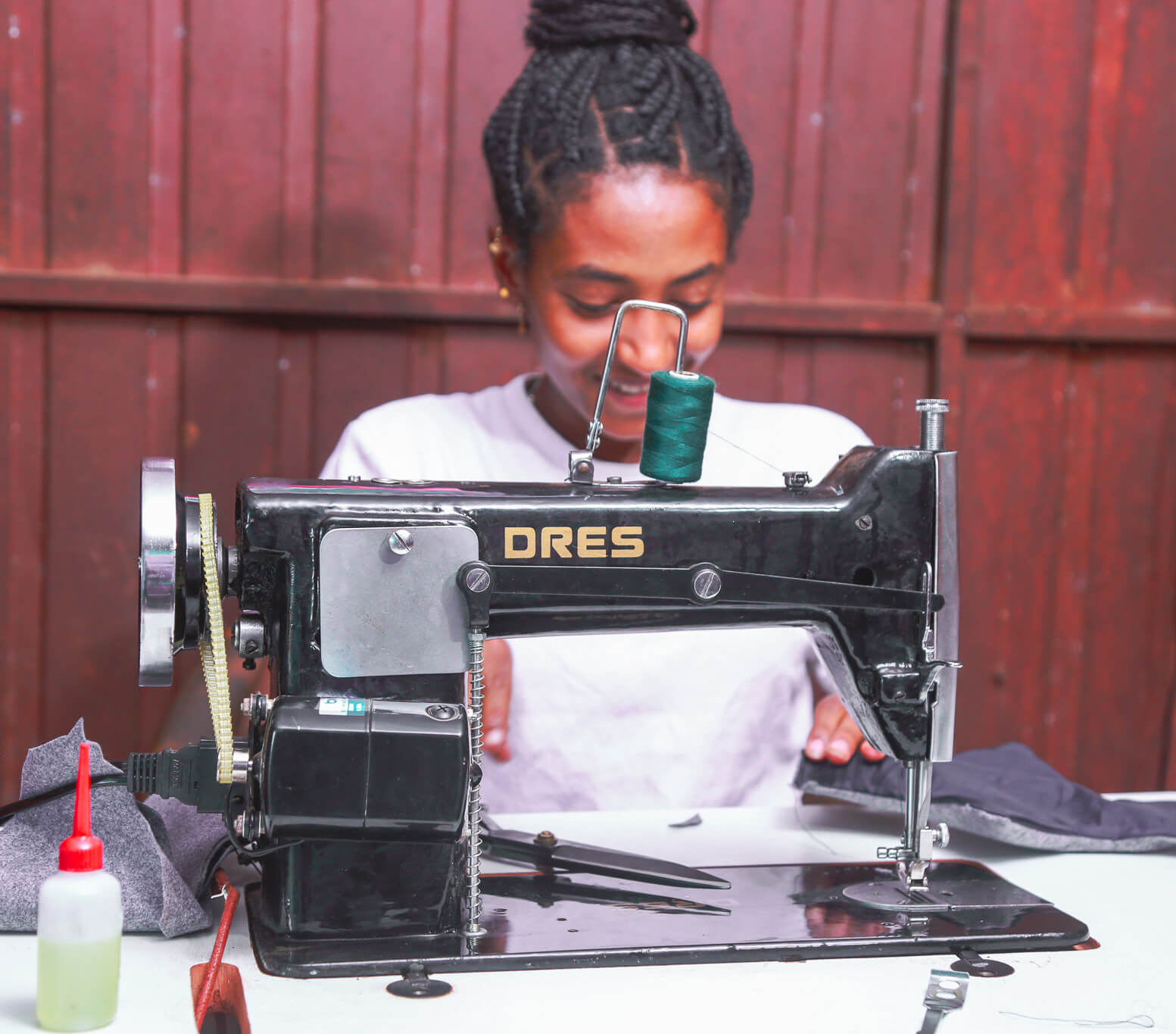 A woman with braided hair smiles while sewing with a black sewing machine on a table, with fabric and tools nearby.