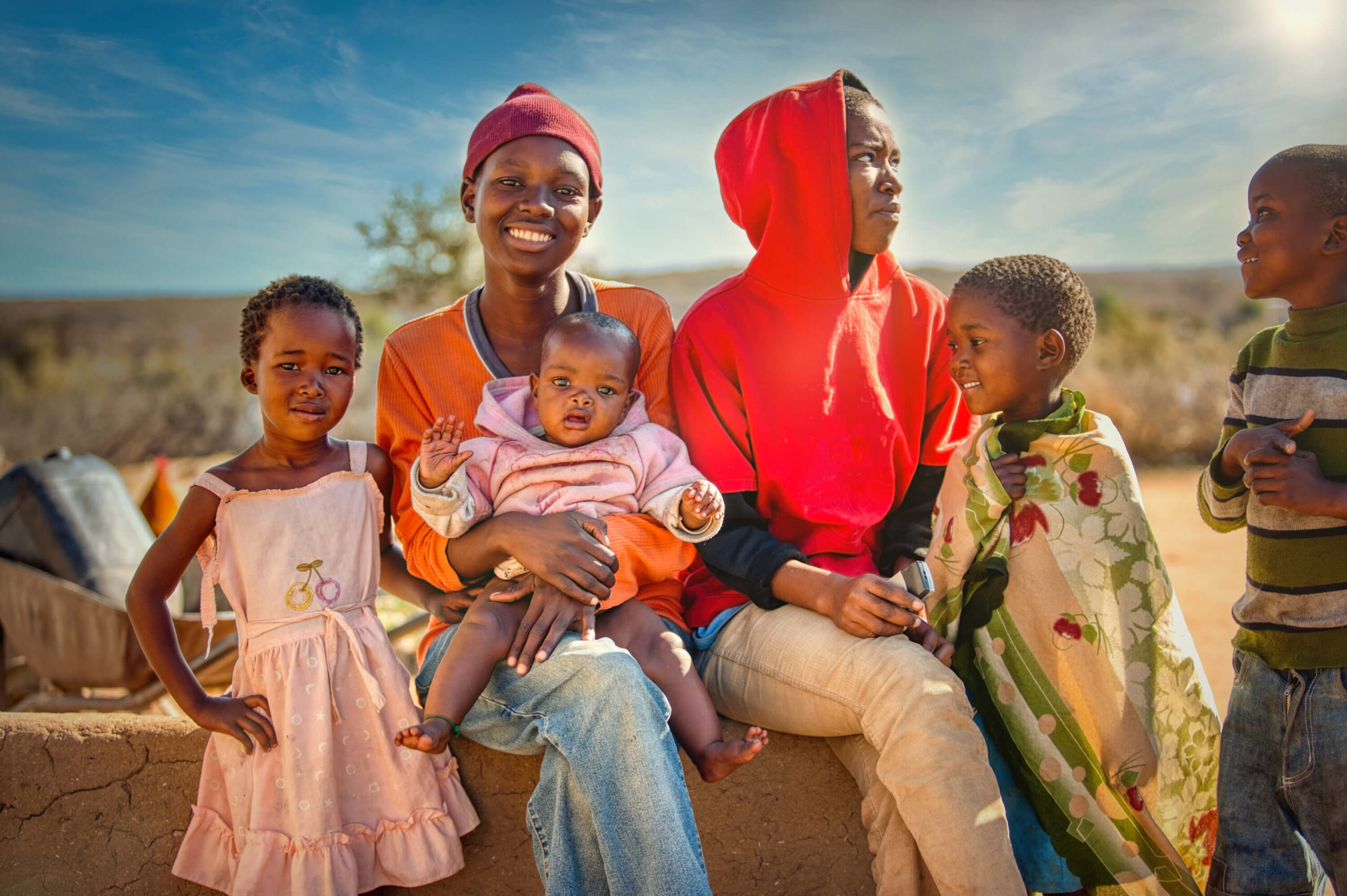 A joyful group of children and a woman, one holding a baby, are sitting together outdoors under a blue sky.