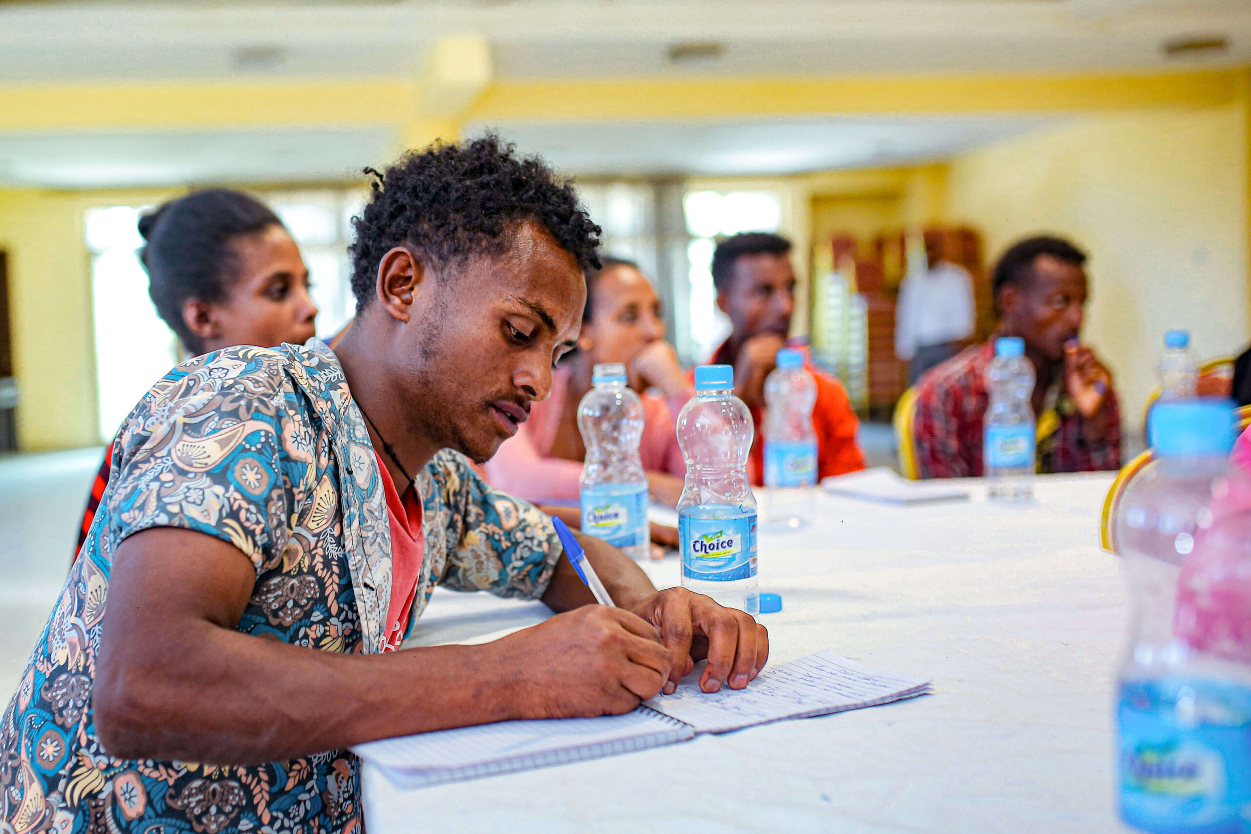 A group of people attend a meeting, focused on a man writing notes at a table with water bottles in the background.