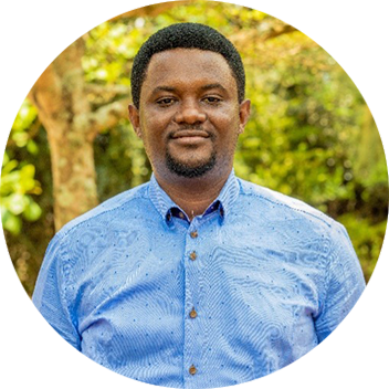 A man with short black hair stands outdoors, smiling in a blue patterned shirt, surrounded by greenery.
