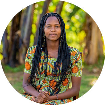 A young woman with long braided hair sits in a garden, wearing a colorful patterned dress with orange accents.
