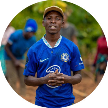 A young man wearing a blue Chelsea jersey and a cap stands with his hands clasped, surrounded by people in a garden.