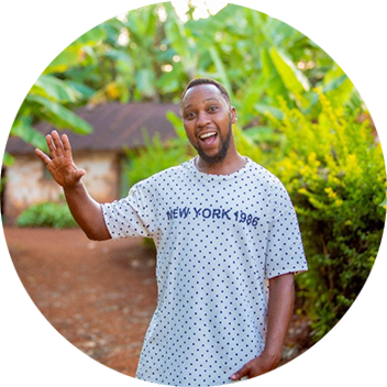 A man in a polka dot shirt smiles and waves in a lush, green outdoor setting.