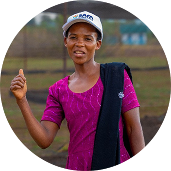 A smiling woman in a pink dress and a cap stands outdoors, gesturing with her hand against a blurred background.
