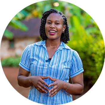 A smiling woman with braided hair stands outdoors, wearing a blue and white striped shirt, gesturing with her hands.