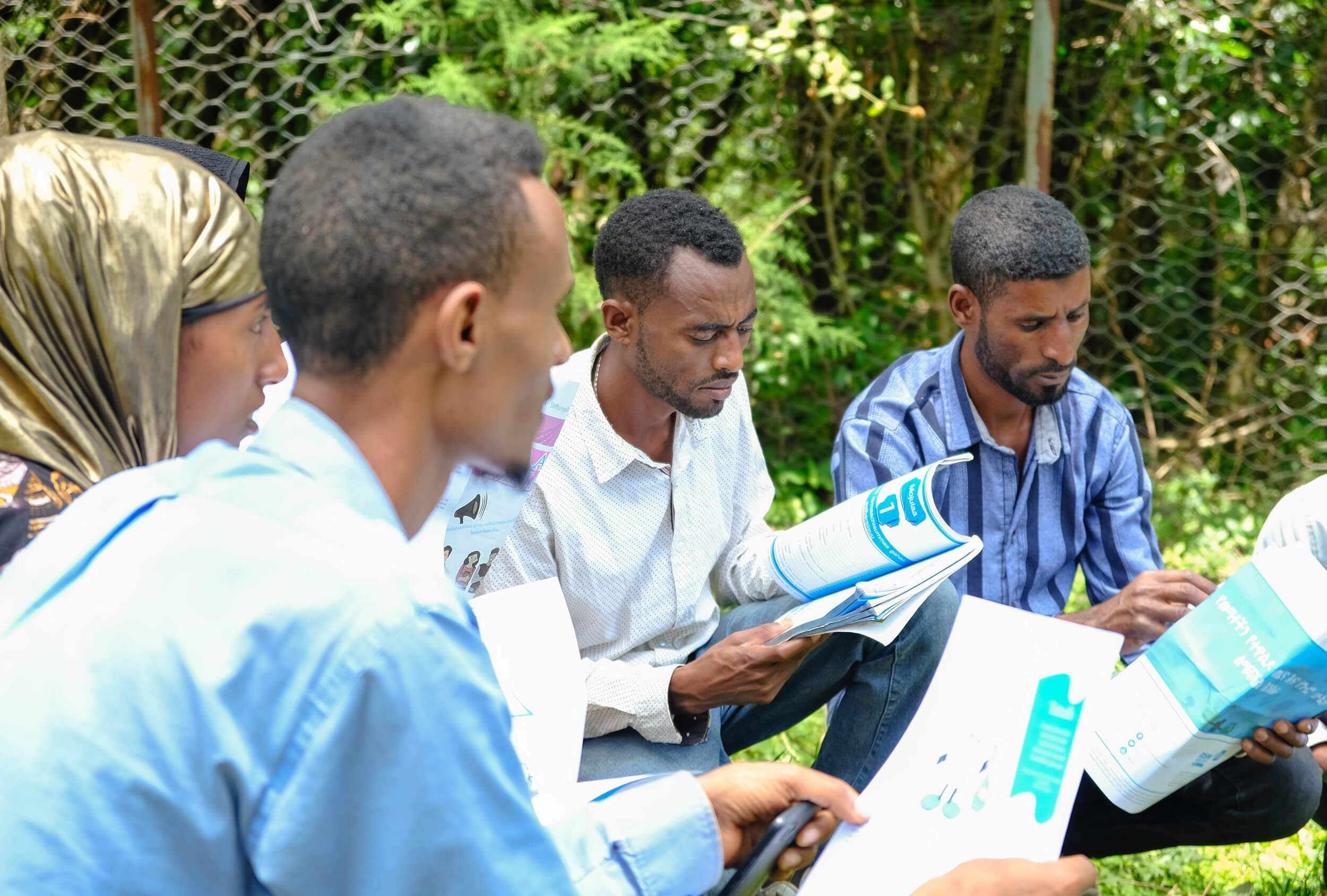 A group of individuals reads pamphlets and brochures while sitting on the grass in a green outdoor setting.