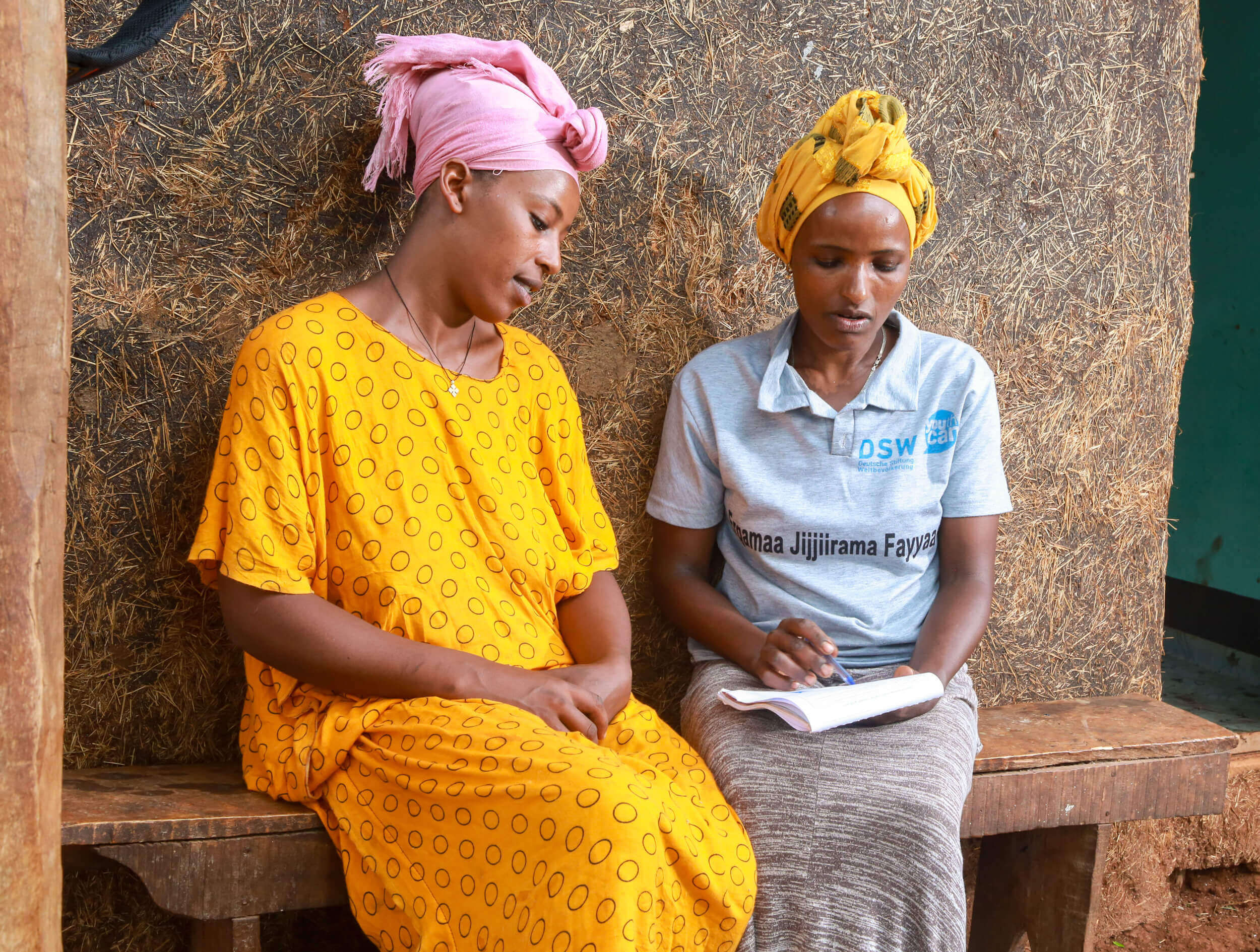Two women sit on a bench, one in a yellow dress with black dots and the other in a gray shirt, discussing and taking notes.