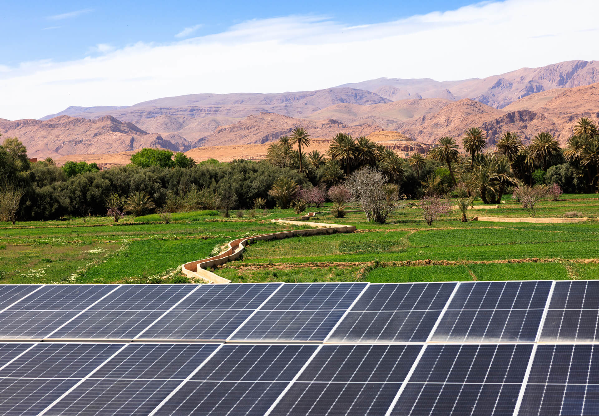 A view of solar panels in the foreground, with lush green fields and mountains in the background under a blue sky.