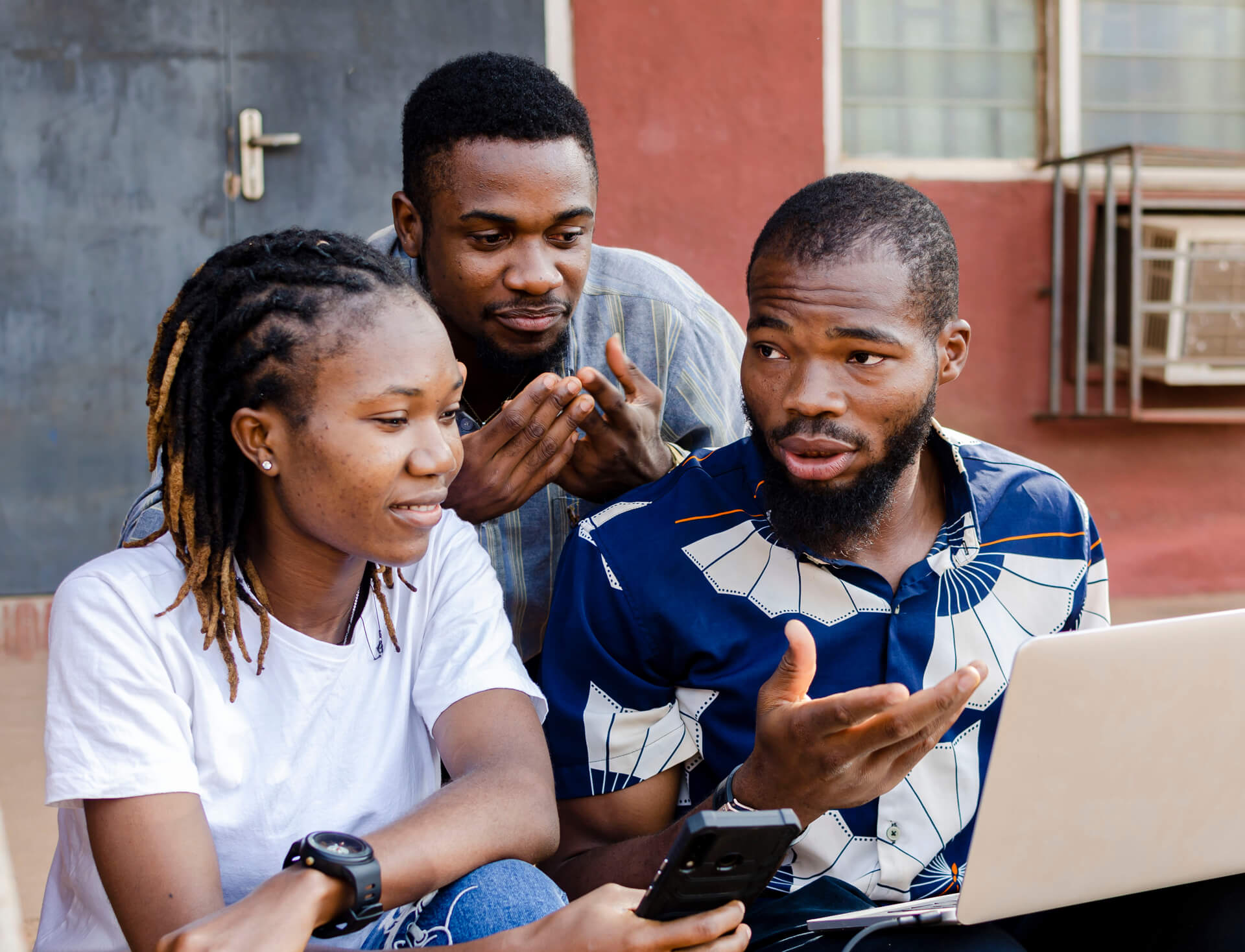 Three people engage in discussion, one on a laptop, another with a phone, while sitting outdoors on a sunny day.