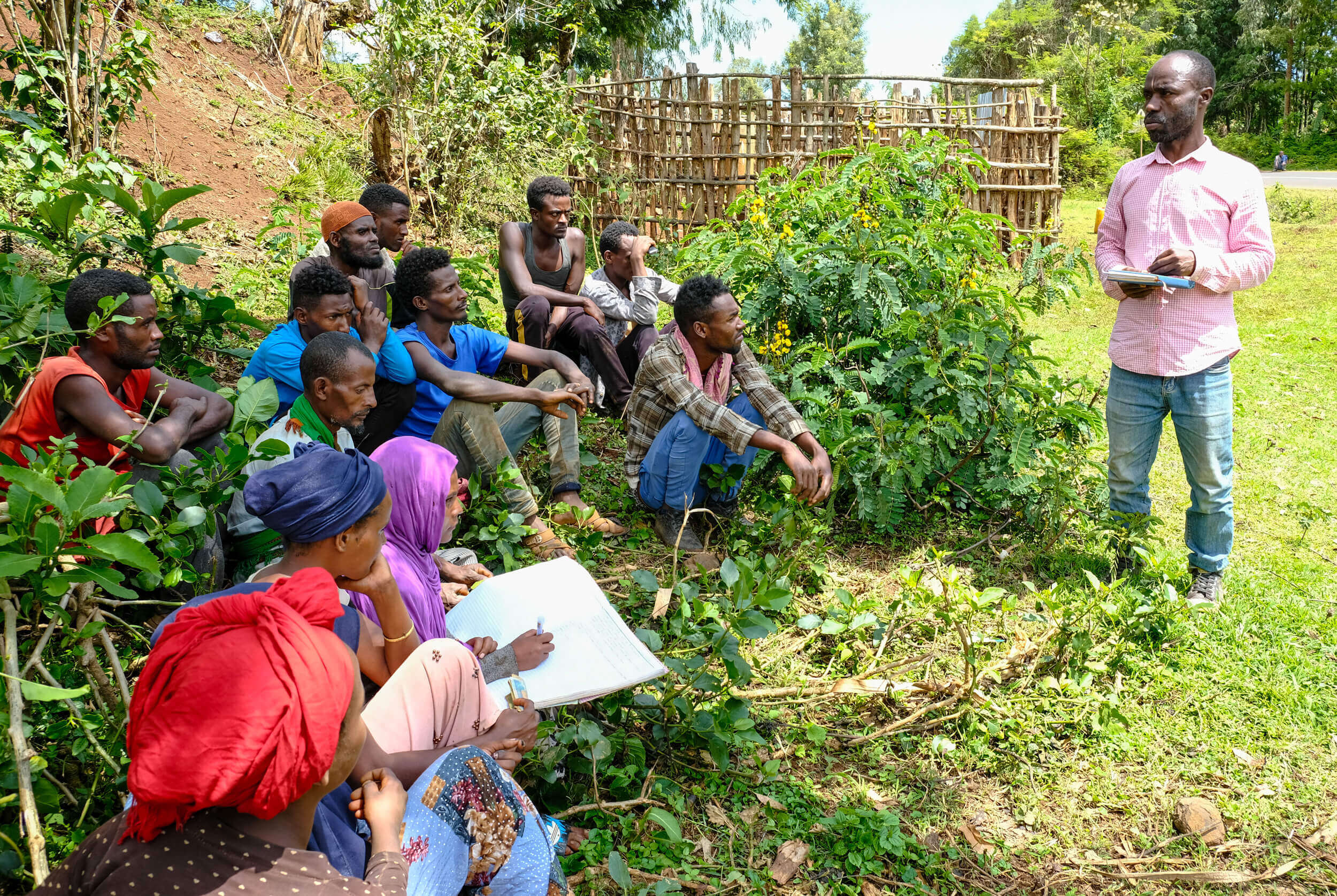 A group of people listen to a speaker outdoors, surrounded by greenery, during a discussion or educational session.