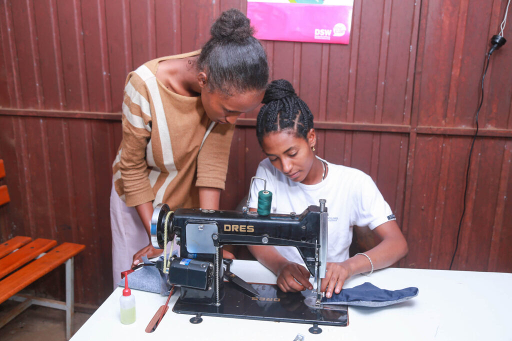 Two women are working together at a sewing machine, with one assisting the other as she sews fabric.