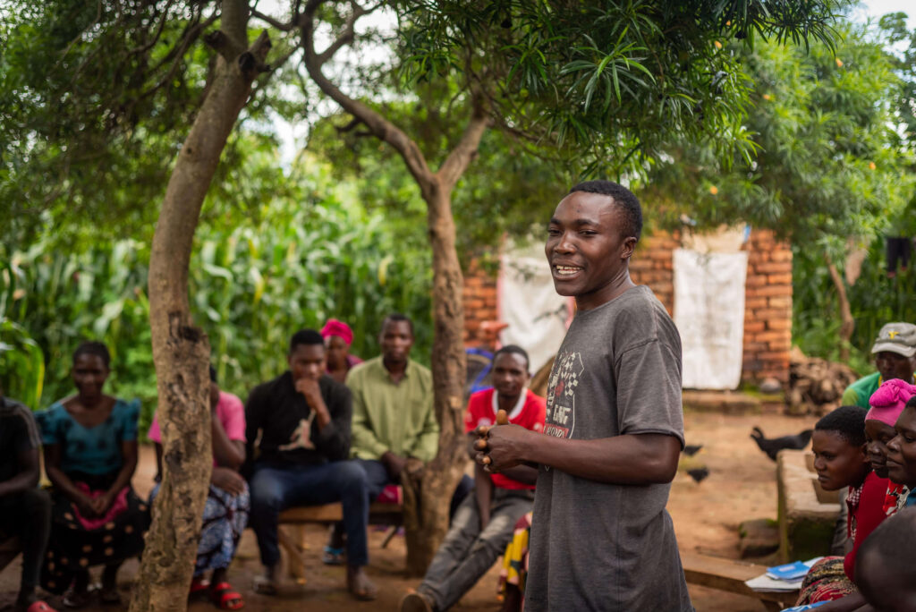 A speaker engages a group seated outdoors, surrounded by trees and greenery, while they attentively listen.