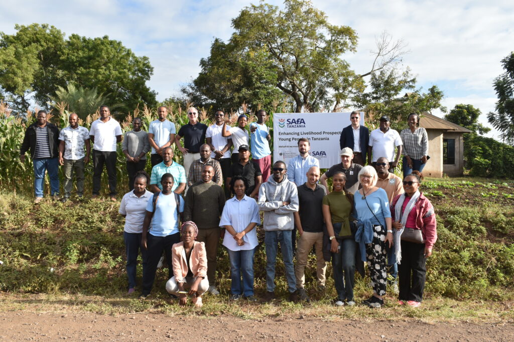 A diverse group of people poses outdoors near a cornfield, with a sign about enhancing livelihood prospects in Tanzania.